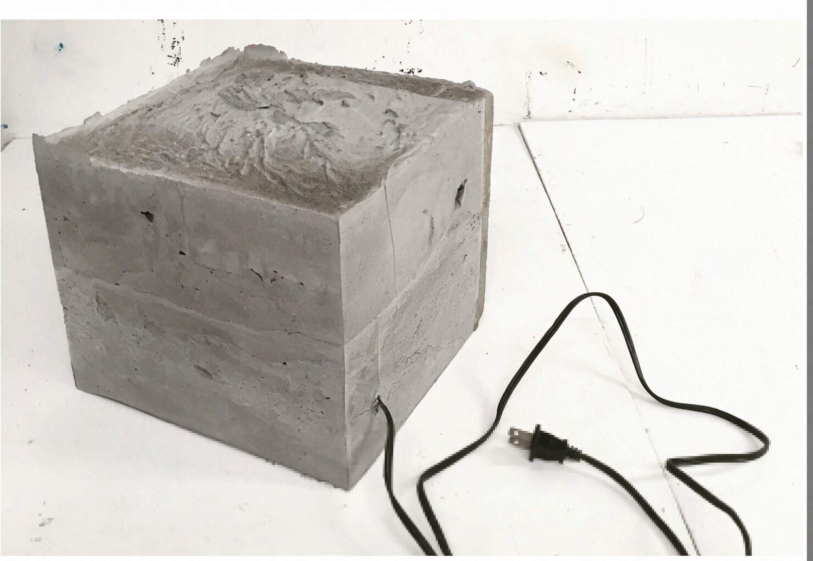 Studio photograph of the existing prototype: a rough, imperfect twelve-inch concrete cube sitting on a white table. A black electrical cord emerges from the lower face of the block and ends in a two-prong plug. The top surface is uneven, with pour marks, voids, and a broken edge.